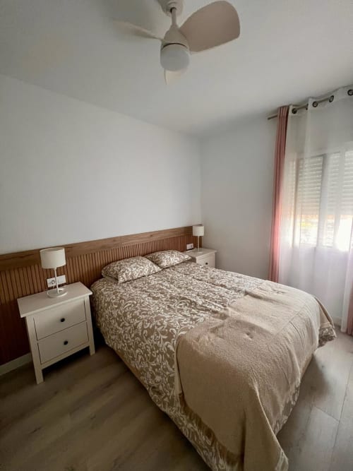 Dormitorio luminoso en apartamento CASA GIL, Águilas: cama de madera, mesilla blanca, ventilador de techo y ventana con vistas al mar con tonos rosados