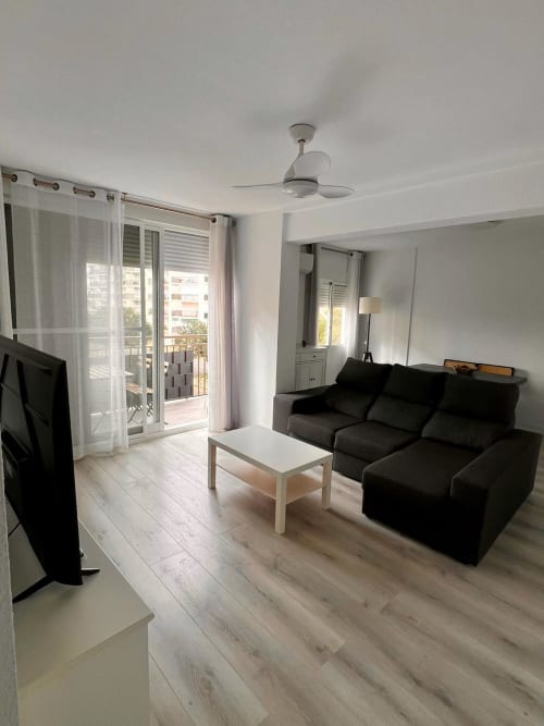 Sala moderna del apartamento Casa Gil en Águilas con sofá negro, balcón con vista al mar, ventilador de techo y piso de madera clara
