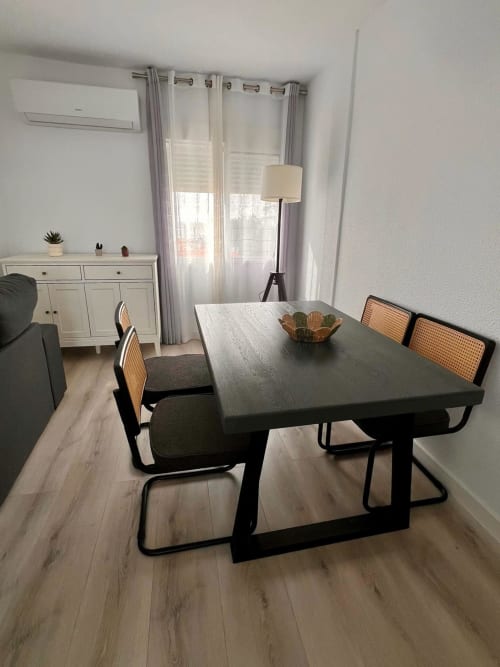 Zona de comedor moderna en apartamento Casa Gil, Águilas con mesa negra, sillas de ratán y vistas al mar a través de cortinas translúcidas
