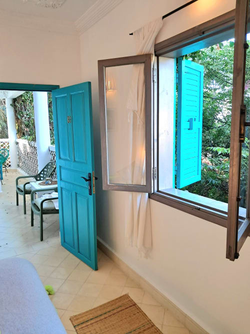 Bright hotel room entrance at Vent Des Dunes with turquoise door, garden view through large window, Essaouira