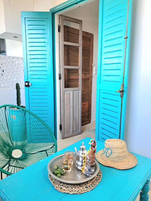Bright turquoise room at Vent Des Dunes hotel in Essaouira with louvered doors, straw hat, and tropical decor on blue table