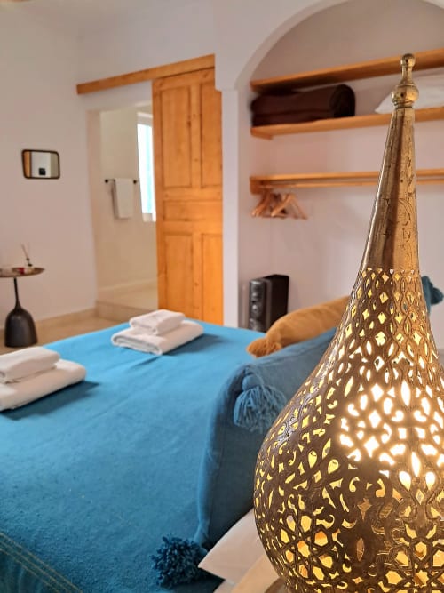 Luxe bedroom at Vent Des Dunes with ornate brass lamp, blue bedding, wooden beams, and arched doorway in Essaouira