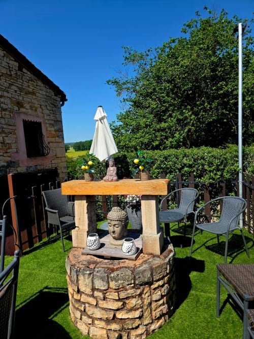 Sonnige Außenbestuhlung bei La Maison de la Colline mit Holztisch, Schirm, Buddhafigur und Landschaftsblick