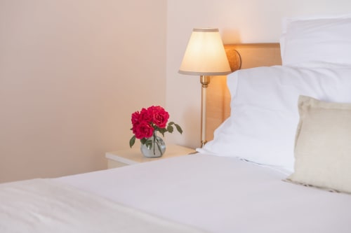 Chambre élégante de l'hôtel Allegria à Bajardo avec draps blancs, lampe de chevet et roses rouges fraîches dans un vase en verre