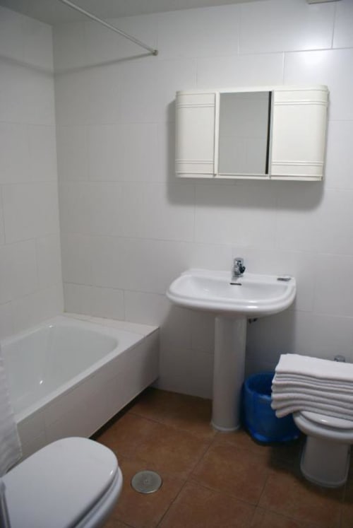 Baño moderno en Alojamientos Botica Rural con azulejos blancos, lavamanos pedestal, bañera, inodoro y espejo botiquín.