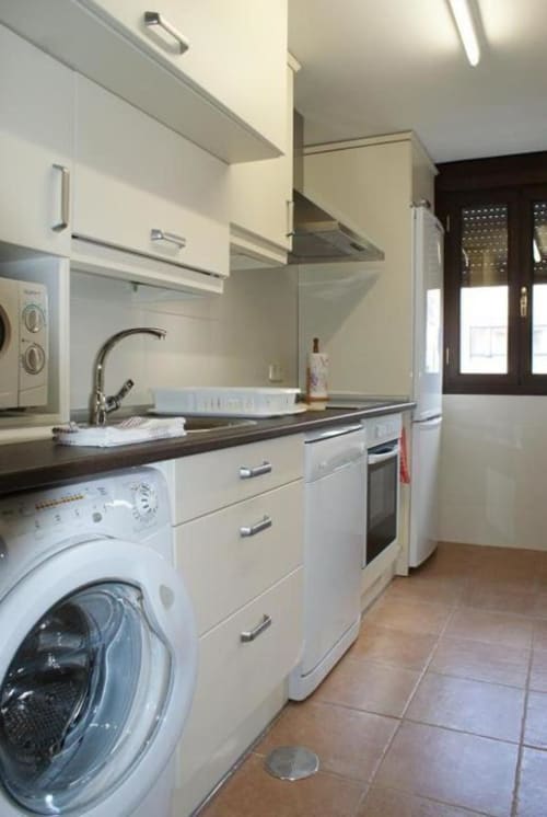 Cocina moderna en apartamento rural Alojamientos Botica con muebles blancos, encimeras oscuras, lavadora y acero inoxidable