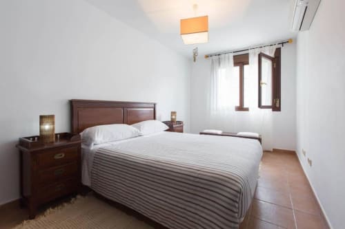 Dormitorio luminoso en apartamento Botica Rural, La Cabrera, con cama de madera, mesita de noche, piso de azulejos y ventana con cortinas.