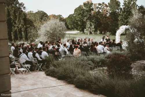Outdoor garden wedding ceremony at Château ERIGOYE in Beychac-et-Caillau, guests seated under olive trees with floral archway