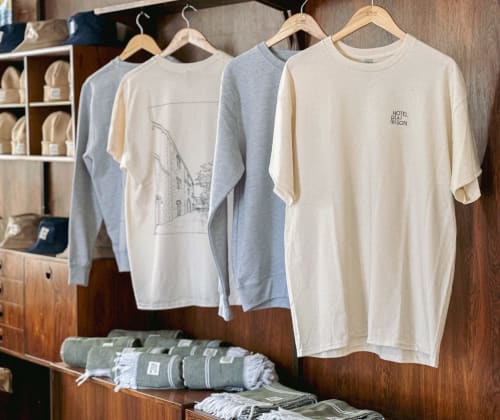 Exhibición de merchandising del Hotel La Prison con camisetas crema y gris en perchas de madera en Béziers