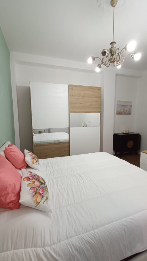 Camera da letto moderna e luminosa a Roma Casa Tua con letto bianco, cuscini rosa, armadio in legno e elegante lampadario