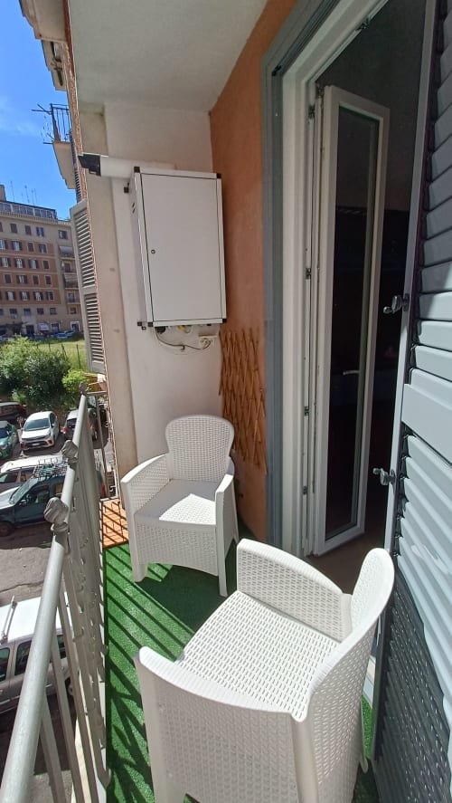 Balcone stretto di un appartamento a Roma con sedie in rattan bianco, erba sintetica verde e vista sulla città presso Roma Casa Tua