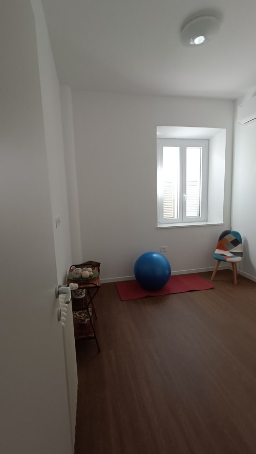 Stanza luminosa e minimalista a Roma Casa Tua con palla fitness blu, tappetino rosso, sedia colorata e finestra con persiane