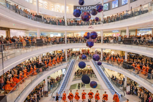 Atrium animé d'une galerie marchande parisienne avec plusieurs étages, foules en tenues oranges, sphères décoratives, escaliers roulants et magasins