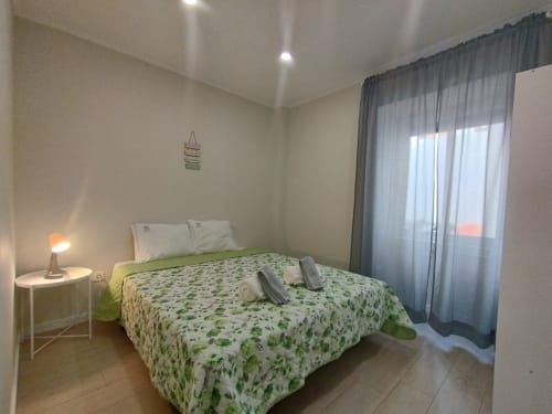 Helles Schlafzimmer in der Furnaka Village Herberge in Lourinhã mit grünem Blattbezug, weißen Kissen, Nachttischlampe und Stadtblick