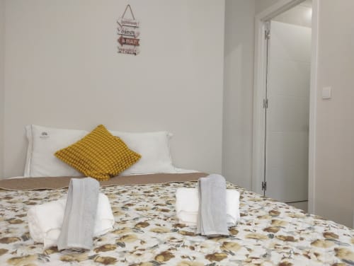 Helles Schlafzimmer in der Furnaka Village Herberge in Lourinhã mit gelbem Kissen, aufgerollten Handtüchern, gemustertem Betttuch und weißer Tür.