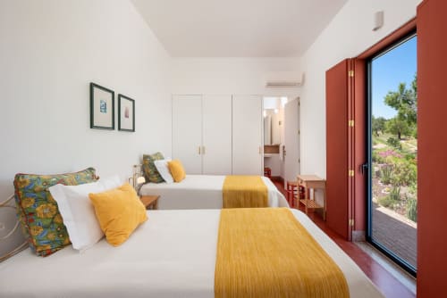 Camas gêmeas com mantas amarelas e almofadas coloridas no quarto luminoso do hotel Tavira com vista para o jardim através das portas vermelhas