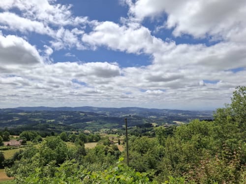 Vue panoramique de la vallée montagne depuis La Maison de Dun avec collines ondulantes, forêts verdoyantes et ciel bleu aux nuages blancs