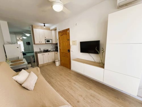 Moderno apartamento estudio en Fuengirola con zona de estar, cocina, TV, suelos de madera y muebles blancos