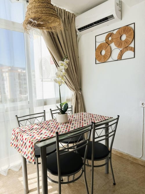 Comedor luminoso en apartamento frente al mar, Fuengirola, con mantel estampado, sillas negras, orquídea y vistas al océano