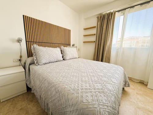 Dormitorio moderno en apartamento frente al mar, Fuengirola. Cama contemporánea con cabecero geométrico, estantes de madera y vistas al mar