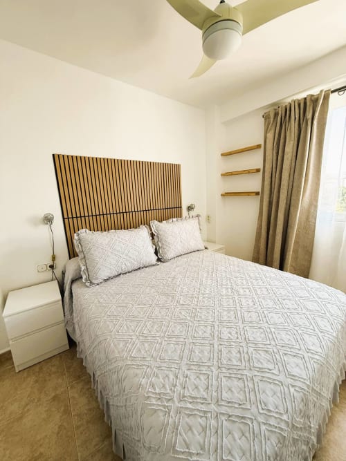 Luminoso dormitorio en apartamento frente al mar en Fuengirola con cabecero rayado, ropa de cama blanca, ventilador de techo y estanterías de madera