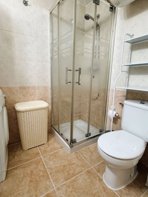 Baño moderno con ducha de cristal, inodoro y cesto de fibra natural en apartamento frente al mar en Fuengirola