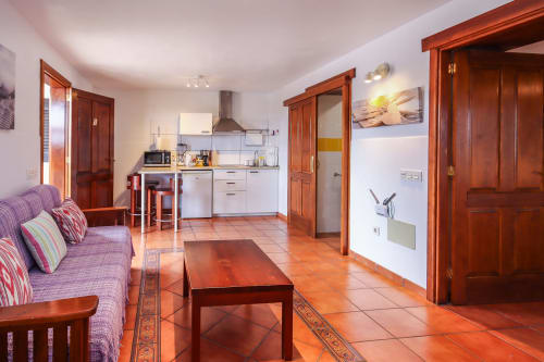 Sala de estar de apartamento diáfano con suelos de barro, puertas de madera, sofá púrpura, cocina americana y mesa comedor