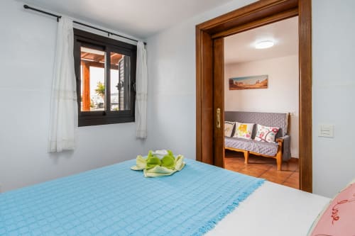 Dormitorio luminoso con cama azul, ventana con vistas a la terraza y puerta de madera hacia la sala en apartamento de Lanzarote