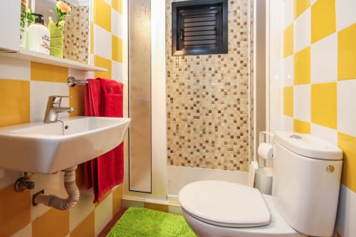 Baño luminoso en amarillo y blanco con lavamanos, inodoro, ducha con azulejos de mosaico y tapete verde en Villanueva Lanzarote