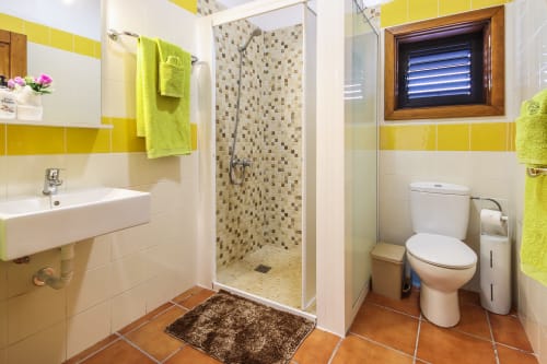 Baño luminoso en amarillo y blanco con ducha de mosaico, lavabo, inodoro y piso de terracota en apartamento Villanueva Lanzarote