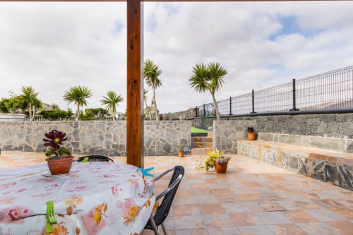 Soleada terraza del apartamento Villanueva en Lanzarote, Mala, con macetas, mesa de comedor, palmeras y muros de piedra