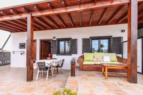 Terraza exterior en Villanueva Lanzarote con pérgola de madera, sofá de mimbre con cojines verdes, mesa de comedor y montaña