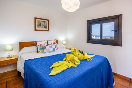 Dormitorio luminoso en Villanueva Lanzarote con edredón azul, muebles de madera, vistas al mar por grandes ventanales y tonos amarillos