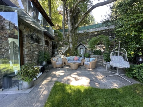 Luxurious courtyard patio with wicker seating, hanging chair, mature trees, and stone buildings at Lake Maggiore retreat in