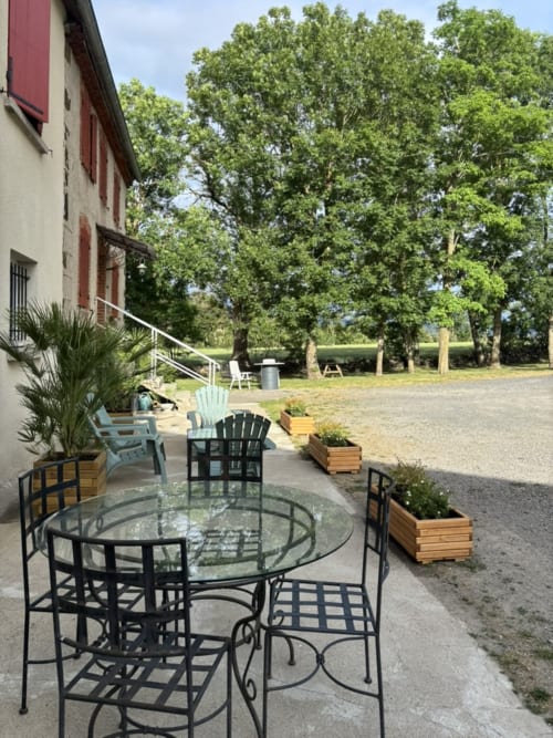 Coin salon extérieur du gîte Mas De Bannayoux avec table ronde en verre, chaises Adirondack et paysage campagnard boisé