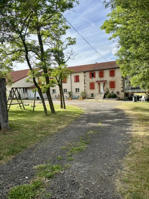 Cour du Mas de Bannayoux avec allée gravillonnée, arbres matures, balançoire et maison de ferme en pierre aux volets rouges à Couteuges