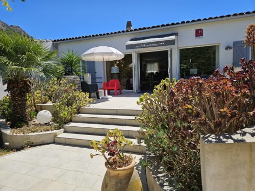 Hotel Les Mouettes terrace in Belgodère with white umbrella, red chair, Mediterranean plants, and stone steps under clear