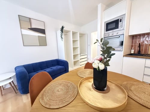 Appartement moderne à Mattbnb Luxembourg avec canapé bleu, salle à manger, table en bois avec fleurs et cuisine blanche