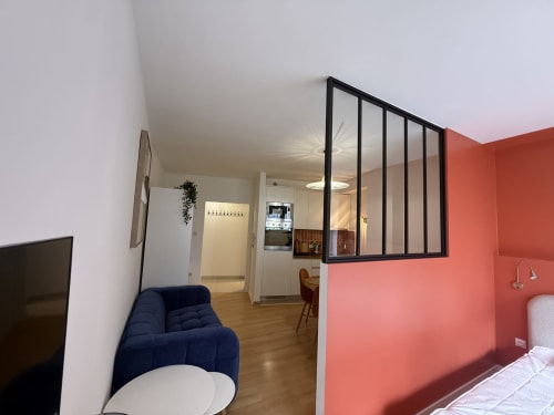 Studio moderne Mattbnb Luxembourg avec canapé bleu, mur d'accent corail et cuisine séparée par une cloison vitrée