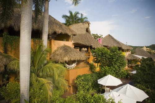 El hotel Ensueño De Fuga en Zihuatanejo cuenta con casitas coloridas con techos de palma, plantas tropicales y sombrillas blancas.