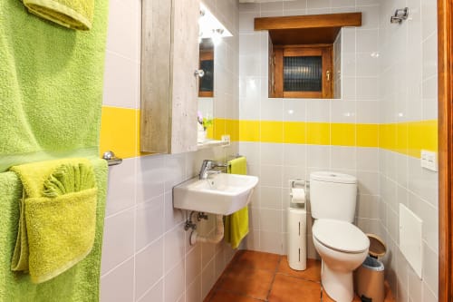 Baño luminoso con azulejos amarillos y blancos, toallas verdes, lavabo, inodoro y piso de terracota en Villanueva Lanzarote