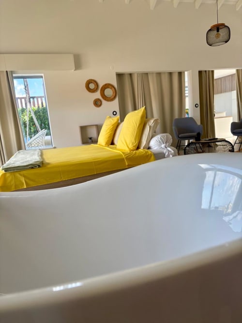 Chambre d'hôtel moderne à Maly Blue Location avec lit jaune, rideaux beiges, miroirs tressés décoratifs et balcon avec vue mer.