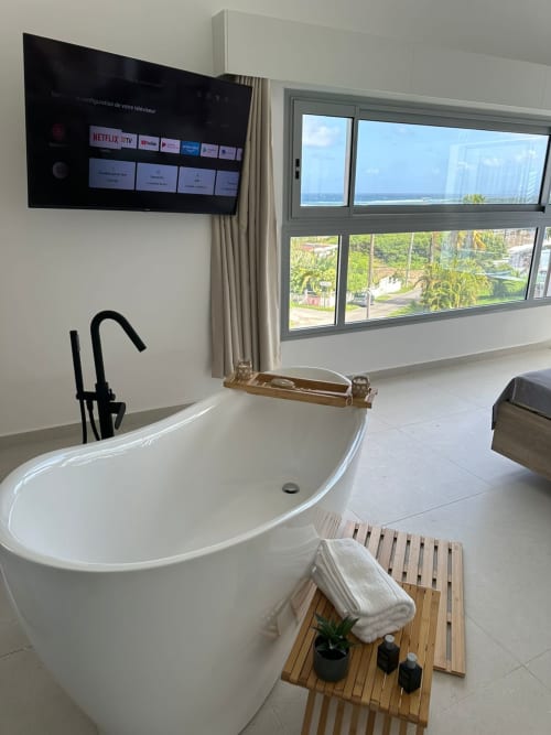 Salle de bain moderne au Maly Blue Location au Moule avec baignoire îlot, TV murale et baies vitrées panoramiques