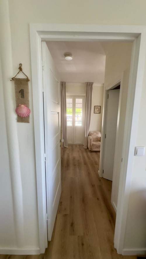 Corredor luminoso da Pink-Shell Family Suite com acesso a portas de varanda e vista para o jardim, piso de madeira e decoração litorânea