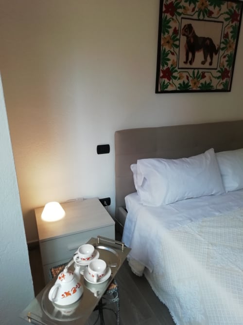 Camera da letto moderna con cuscini bianchi, servizio di tè sul comodino e quadro con ritratto di cane presso House Live Express a Quartu