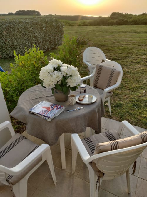 Terrasse extérieure de l'appartement Locmaria Belle Île avec chaises blanches, table ronde, fleurs blanches, magazine et coucher de soleil