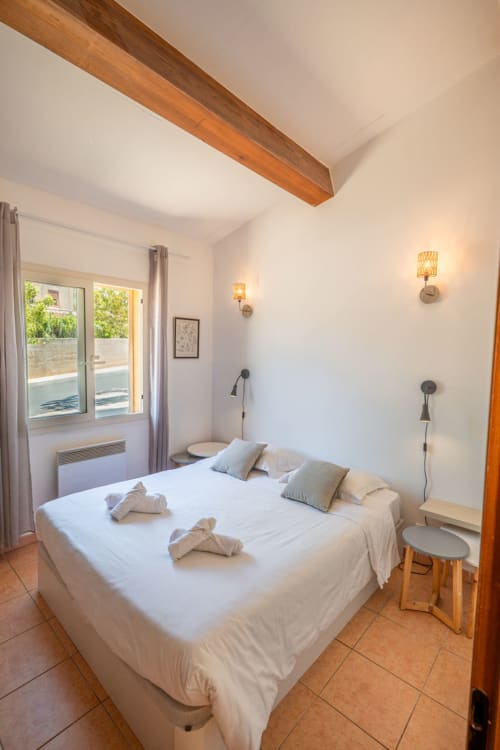 Chambre lumineuse de la villa de Propriano avec lit queen-size, plafond à poutres apparentes, portes donnant sur jardin, appliques murales et carrelage en terre cuite