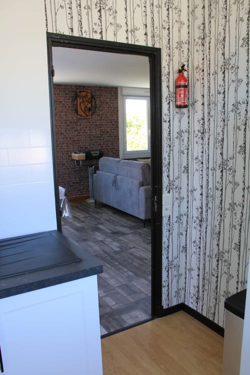 Entrée du gîte Le Grand Bain à La Pause Landaise, montrant le séjour avec fauteuil bleu, mur en brique et vue sur le jardin