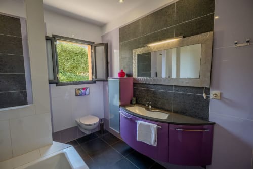 Salle de bains moderne avec meuble de rangement violet, carrelage foncé, grand miroir éclairé et fenêtre donnant sur le jardin à la Résidence
