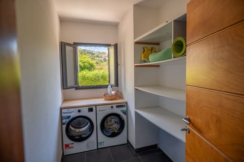 Salle de lavage moderne avec machines à laver côte à côte et sèche-linge, armoires en bois, fenêtre ouverte avec vue sur les montagnes